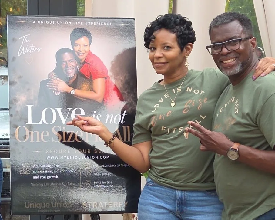 Couple wearing matching olive Love Is Not One Size Fits All shirts beside an event sign